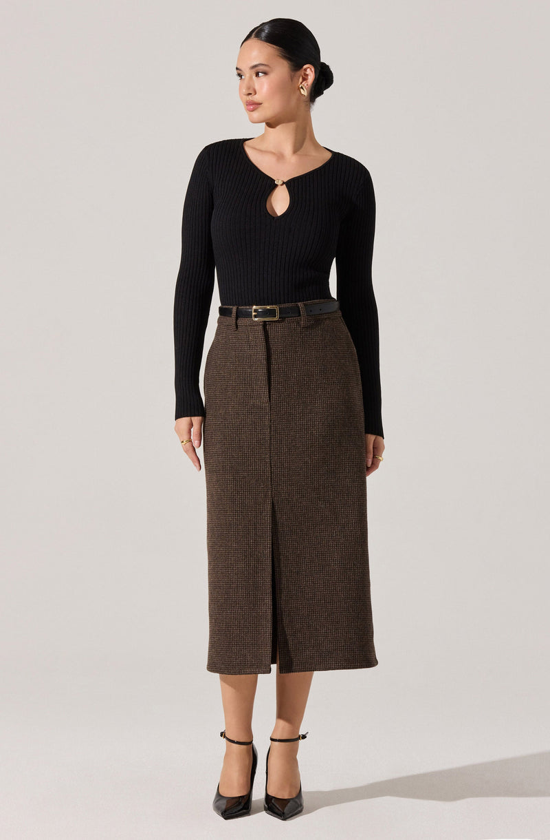 A woman is wearing a fitted, long-sleeved, black ribbed sweater with a small keyhole and metal ring detail at the neckline. She pairs it with a high-waisted, brown, checkered midi skirt that has a front slit and a black belt with a gold buckle. She completes the outfit with black pointed-toe high heels with ankle straps.