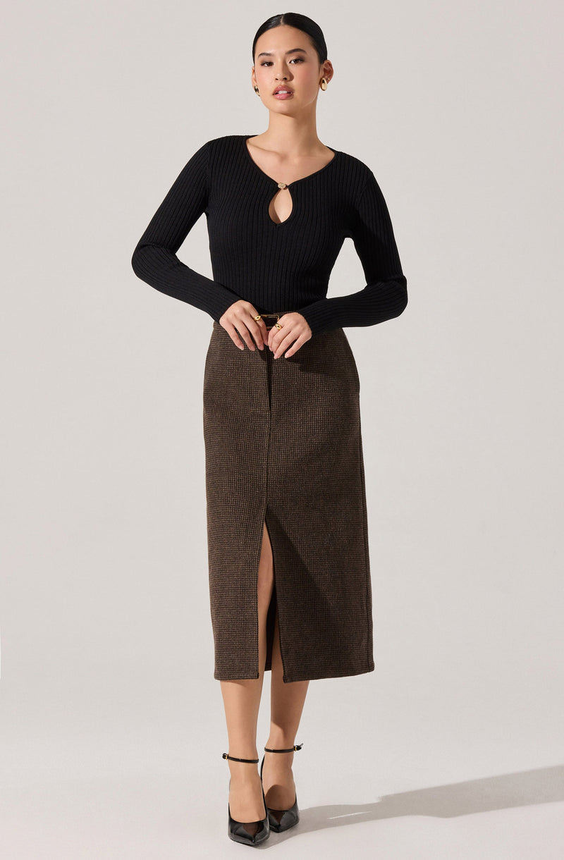 A woman is wearing a black, long-sleeved, ribbed knit top with a keyhole neckline and a small metal ring detail. She pairs it with a high-waisted, brown midi skirt that features a front slit and a subtle checkered pattern. She completes her outfit with black ankle-strap heels.