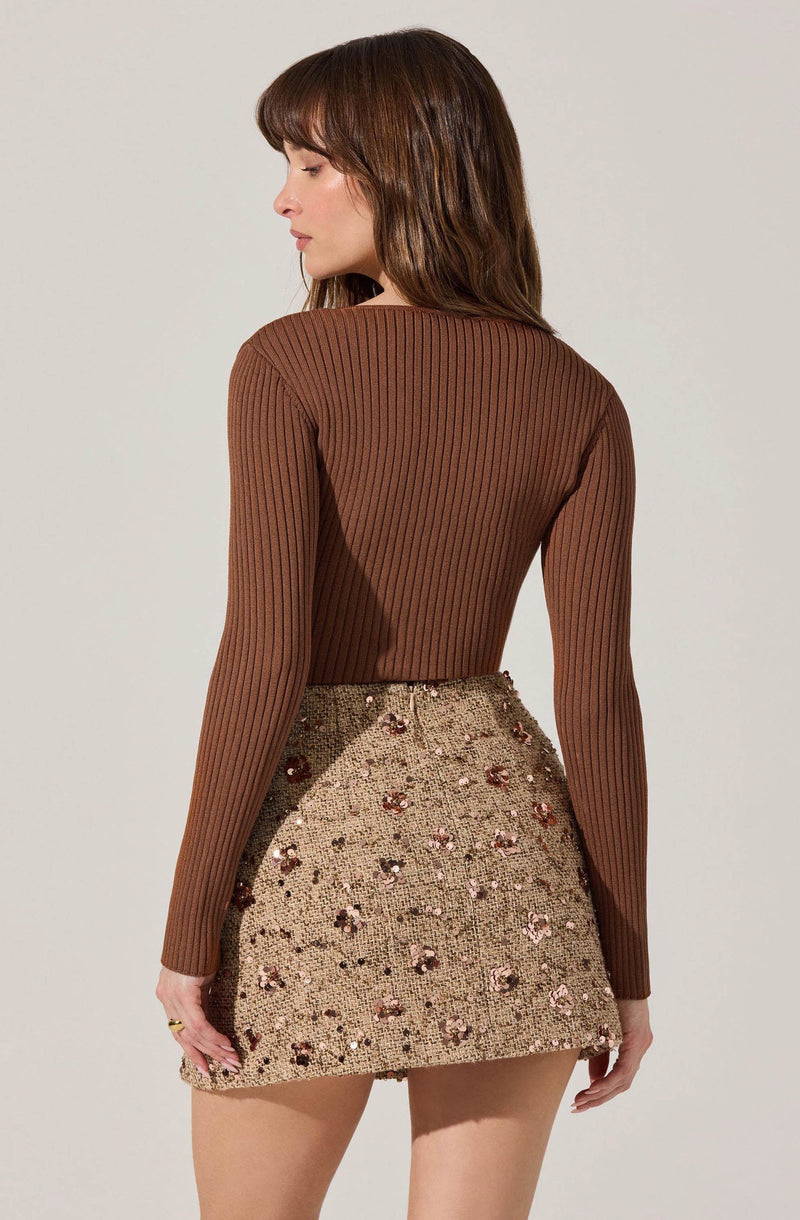 A woman is wearing a fitted, long-sleeved, ribbed brown sweater and a high-waisted, short skirt embellished with sequins and floral embroidery in shades of pink, brown, and beige.