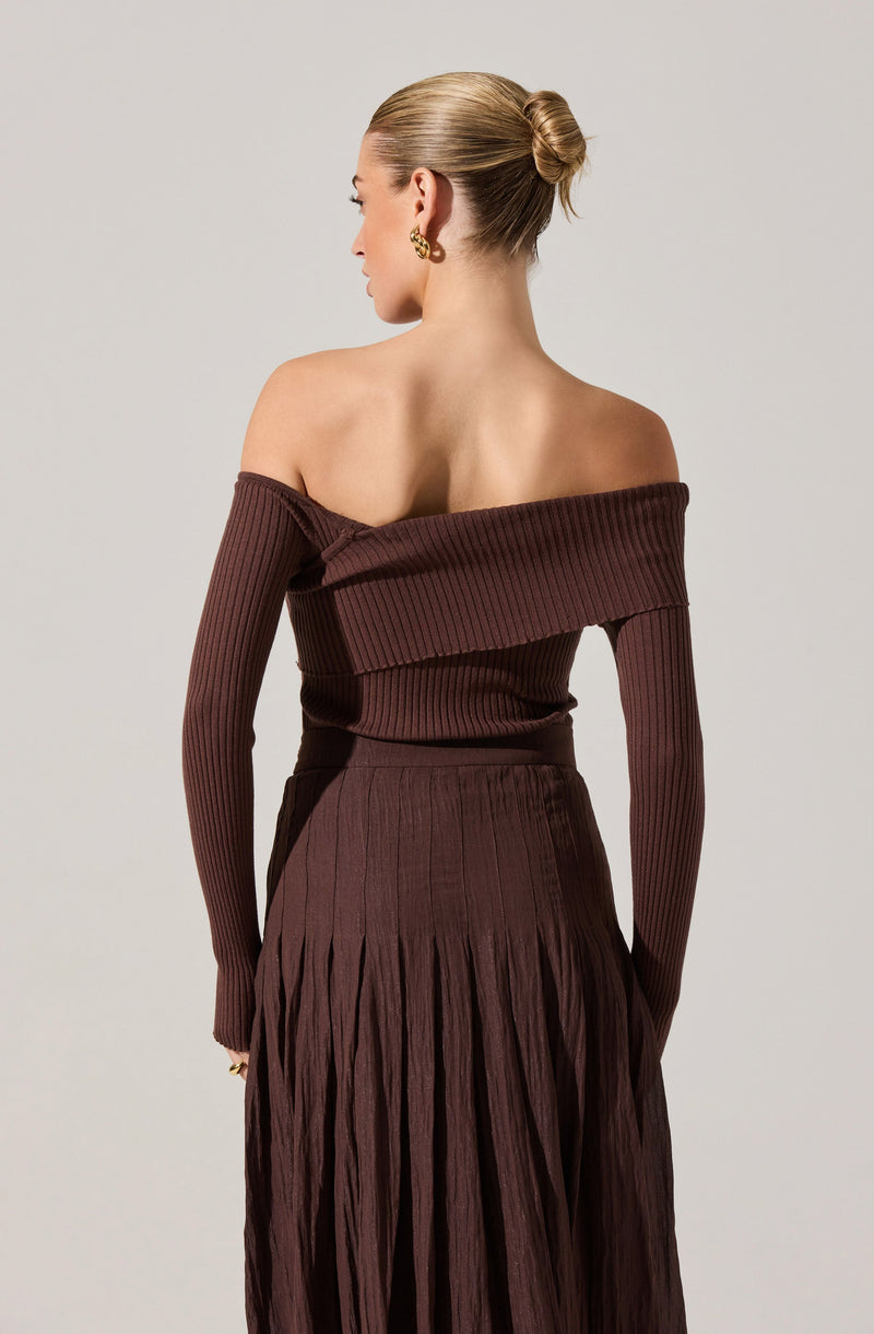 A woman is wearing a coordinating set in a deep brown color. The off-the-shoulder top has long sleeves and features a wide, folded-over neckline with a ribbed texture. The high-waisted skirt is also ribbed at the waist and transitions into pleated fabric that extends to the floor. She accessorizes with gold earrings and a ring, and her hair is styled in a sleek bun.