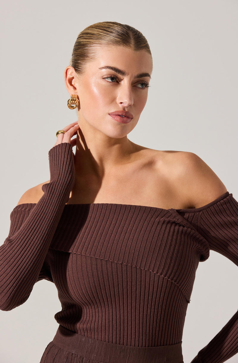 A woman is wearing an off-the-shoulder, long-sleeved, ribbed sweater in a deep brown color. The sweater has a wide neckline that exposes her shoulders and collarbone, and the ribbed fabric features vertical lines. She is accessorized with gold hoop earrings and a gold ring. Her hair is styled in a sleek, compressed look pulled back from her face.