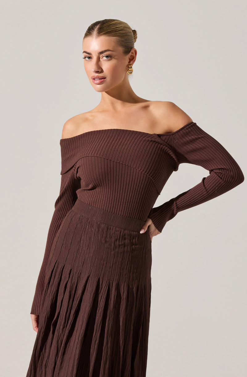 A woman is wearing a brown, off-the-shoulder, ribbed knit top with long sleeves that drape over her arms. She pairs it with a matching high-waisted, pleated skirt in the same brown color that extends down past her hips. The outfit features a textured, vertically pleated design throughout.