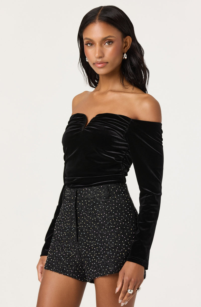 A woman is wearing an off-the-shoulder black velvet top with long sleeves and a ruched front. She pairs it with high-waisted black shorts adorned with multicolored rhinestones or beads, creating a sparkly, embellished look.