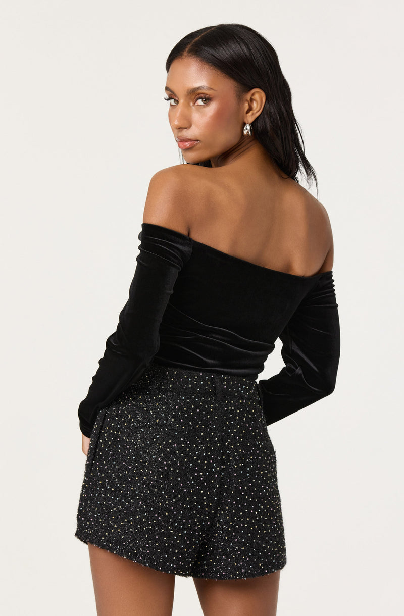 A woman is wearing a black off-the-shoulder long-sleeve top made of a velvety fabric, paired with high-waisted black shorts adorned with small sparkly embellishments. She is also wearing a pair of dangling earrings.