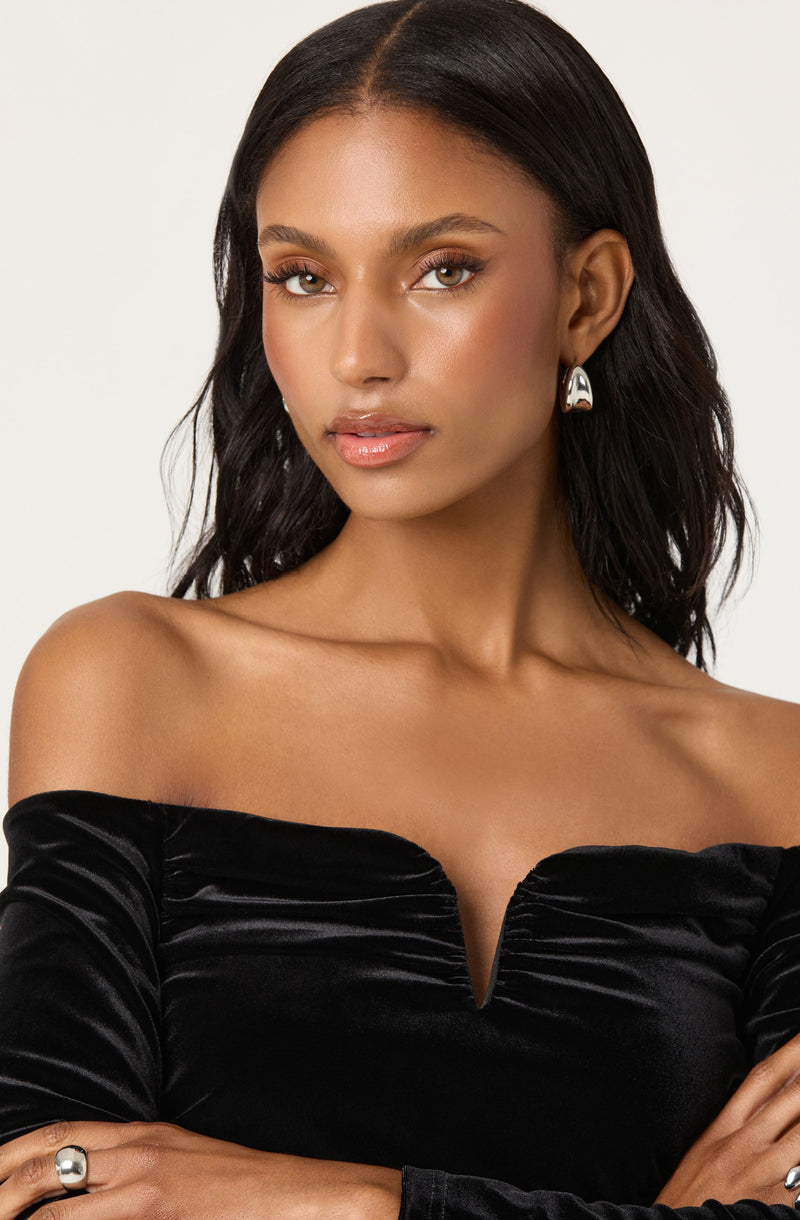 A woman is wearing an off-the-shoulder black velvet top with a slight V-shaped neckline. She has on silver earrings and a matching silver ring on her right hand.