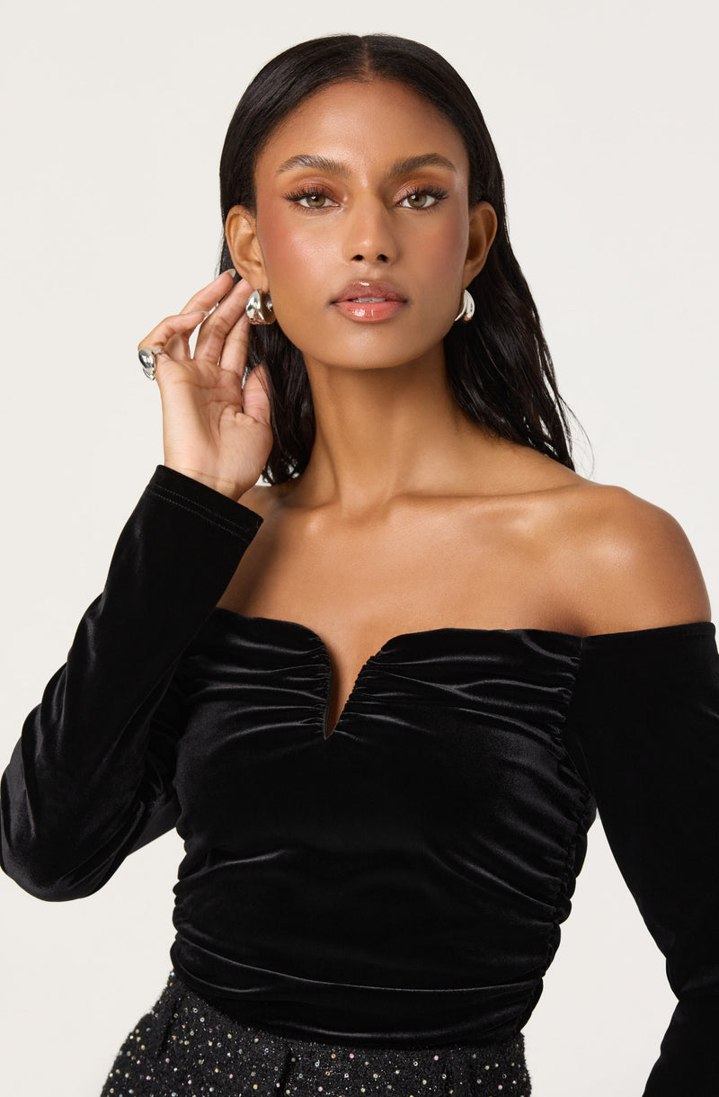 A woman is wearing an off-the-shoulder black velvet top with a ruched design at the front. She accessorizes with large pearl earrings and a ring on her right hand.