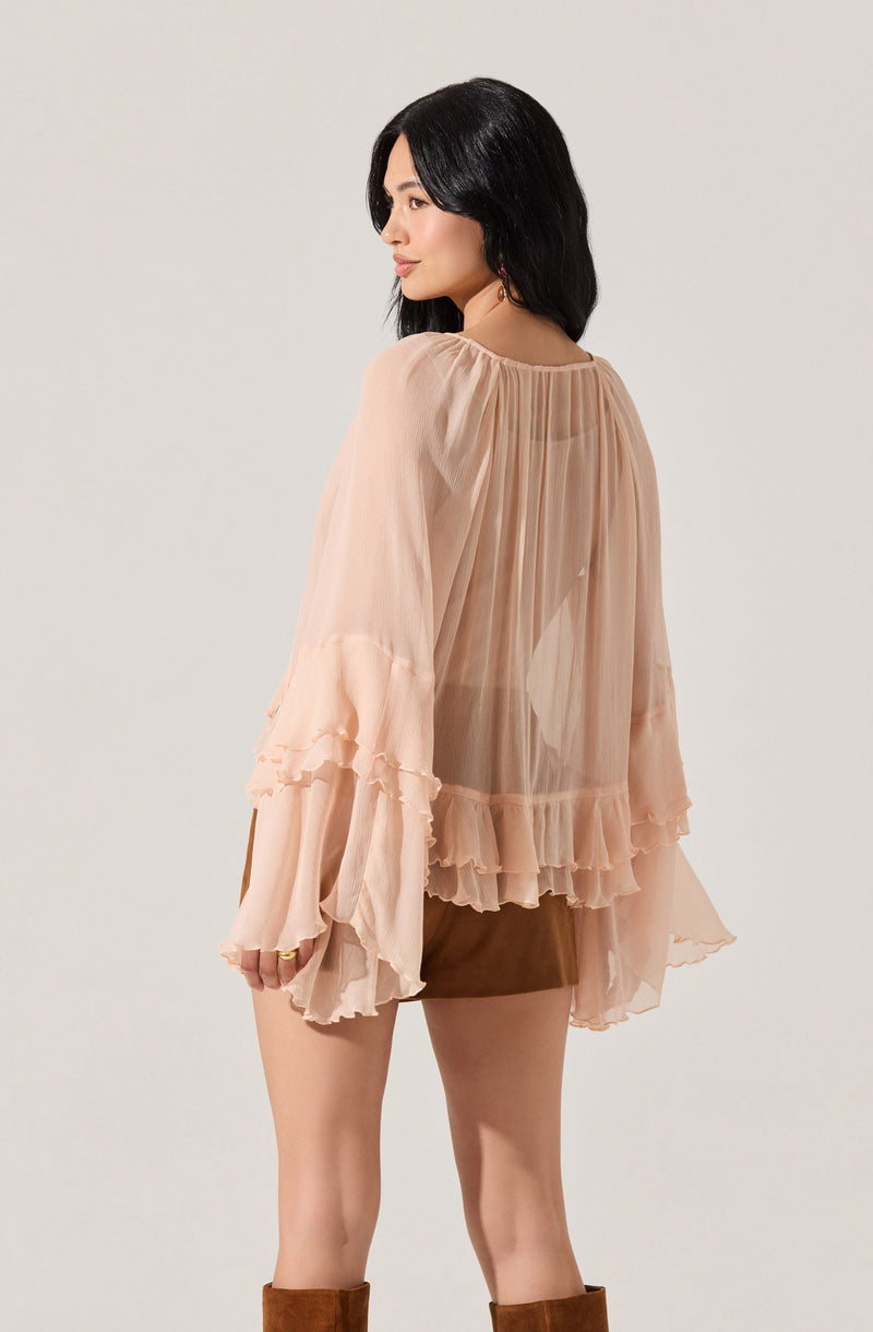 A woman is wearing a light, sheer, peach-colored blouse with long, loose sleeves that have ruffled, layered cuffs. The blouse features vertical pleats running down the back and ruffled hems at the bottom. She is also wearing a short brown skirt and brown knee-high boots.