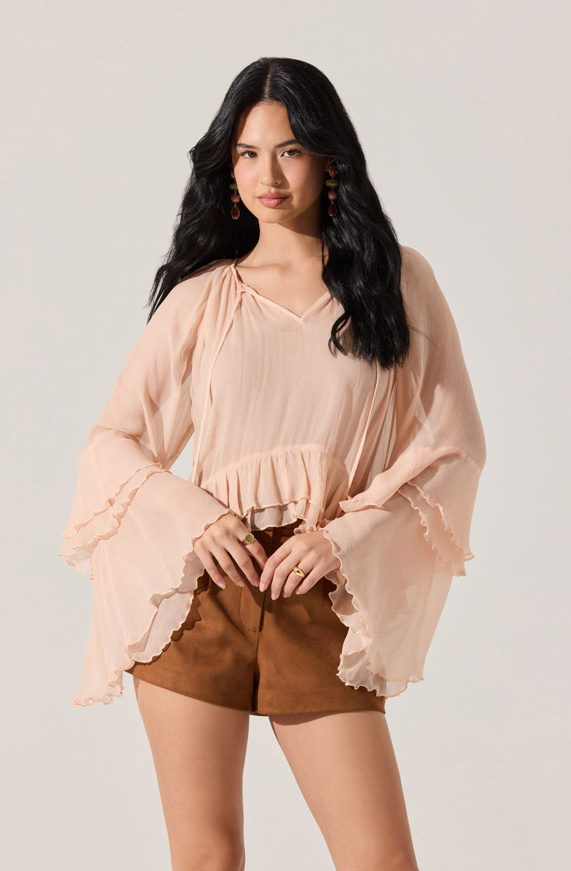 A woman is wearing a light peach, sheer, flowy blouse with voluminous, ruffled, bell sleeves and a ruffled hem. She pairs the blouse with brown shorts.
