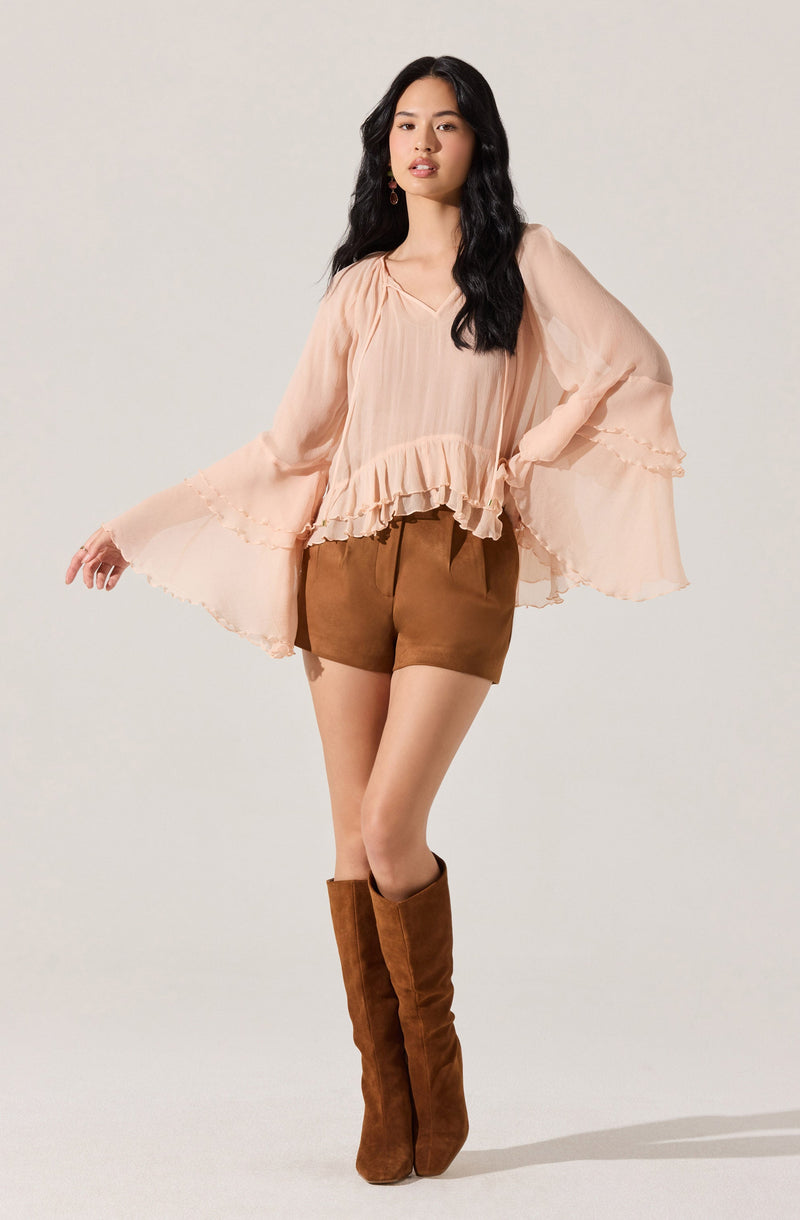 A woman is wearing a loose, flowing, peach-colored blouse with ruffled details at the bottom hem and around the wide, billowy sleeves. She pairs it with brown shorts and knee-high brown suede boots.