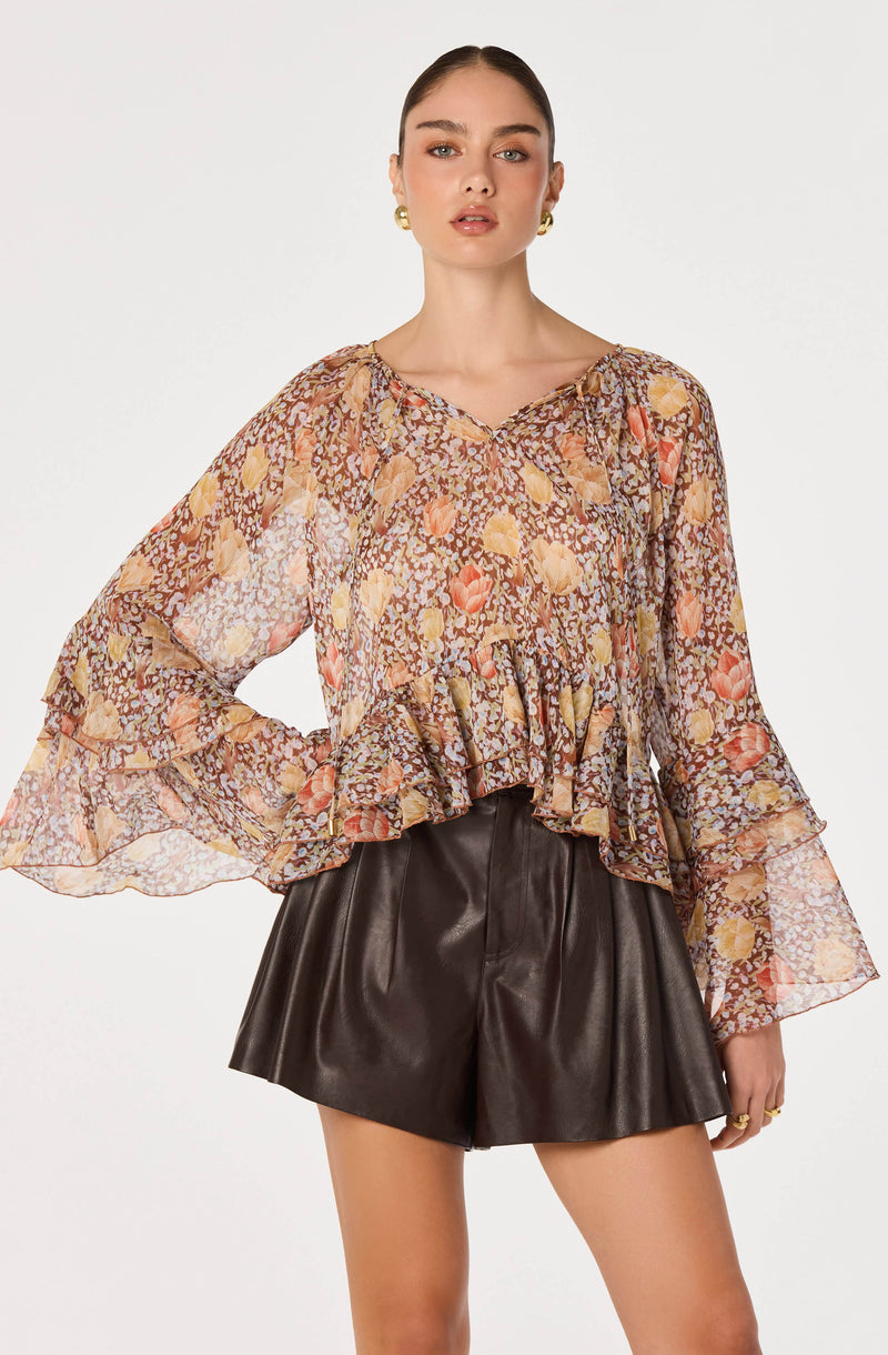 Woman wearing a sheer, floral printed blouse with ruffled hem and wide bell sleeves, paired with black leather shorts.