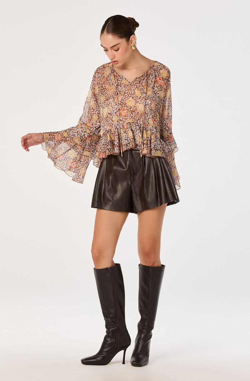 Woman wearing a flowy, floral-patterned blouse, black leather shorts, and tall black heeled boots.