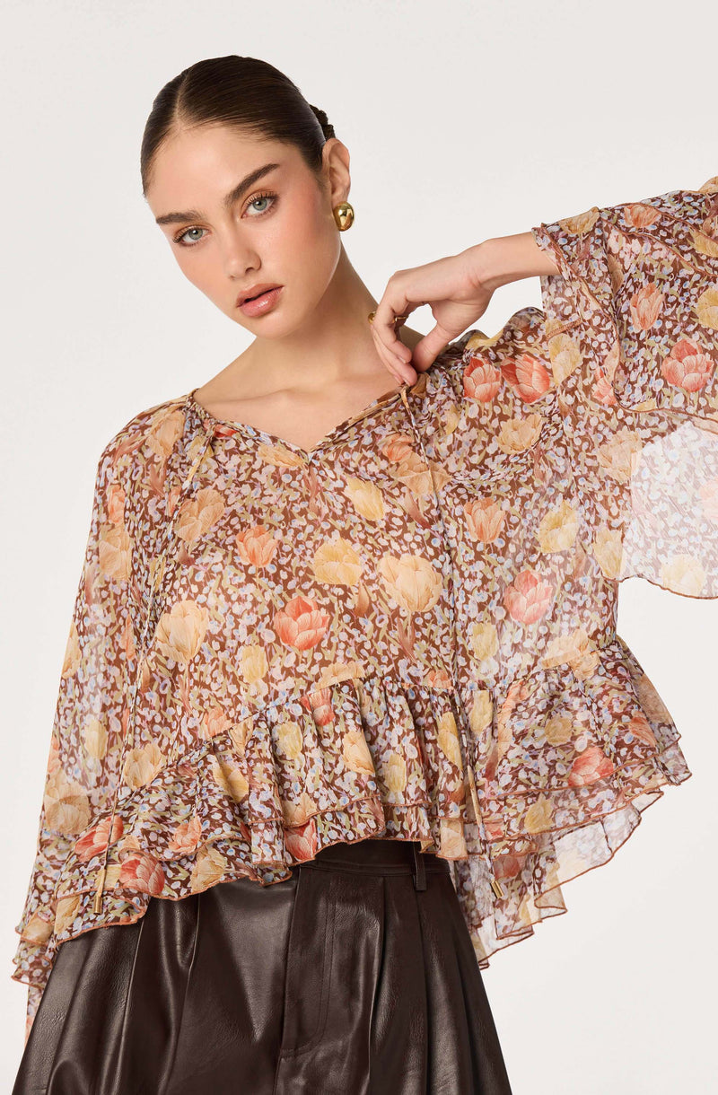 Woman wearing a sheer, floral-patterned blouse with ruffled hem and wide sleeves.