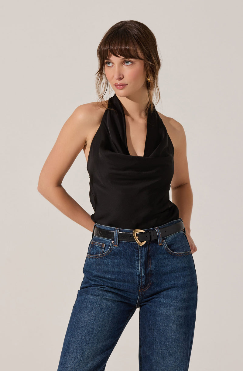 A woman is wearing a sleeveless black satin top with a draped cowl neckline, paired with high-waisted blue jeans that have a classic fit. She has accessorized with a black belt featuring a gold buckle and small gold hoop earrings.