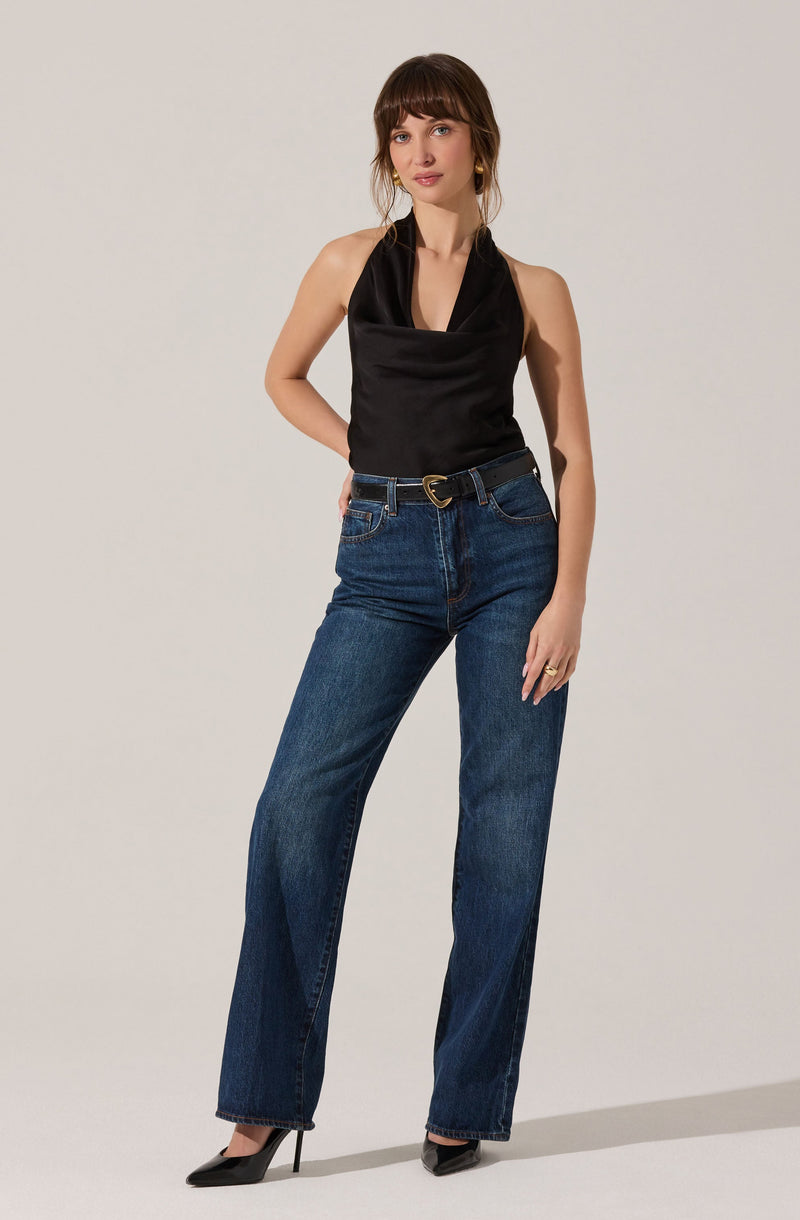 A woman is wearing a sleeveless black top with a cowl neckline, paired with high-waisted blue jeans. She has on black pointed-toe high heels.