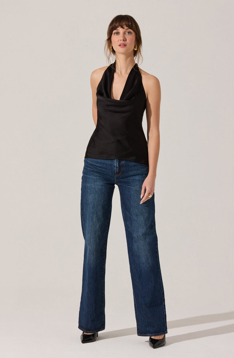 A woman is wearing a sleeveless black satin top with a draped cowl neckline, paired with high-waisted, straight-leg blue jeans and black pointed-toe shoes. She has shoulder-length brown hair with bangs and is standing against a plain, light-colored background.
