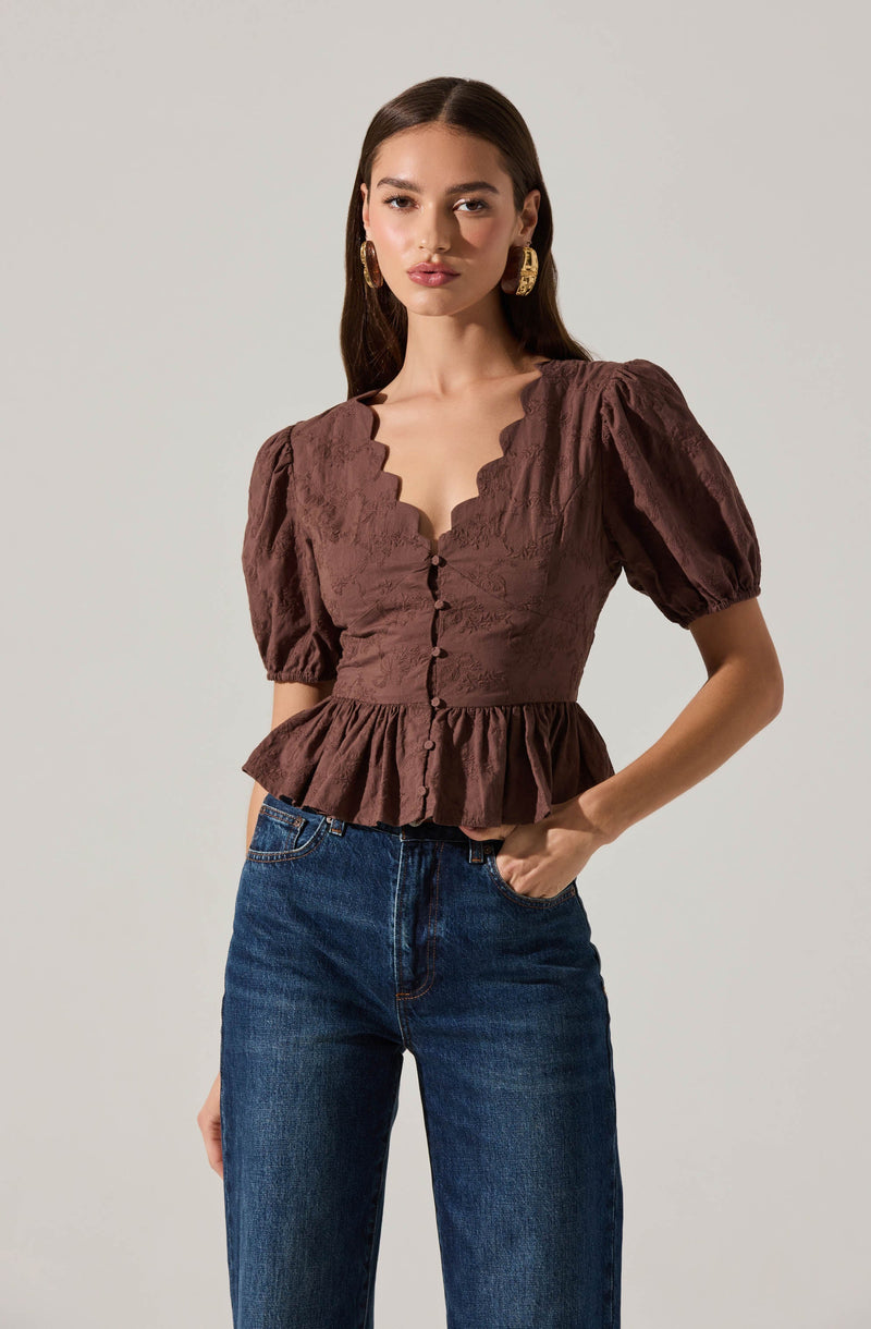 A woman is wearing a brown blouse with a scalloped neckline and puffed short sleeves. The blouse features decorative buttons down the front, a fitted waist, and a ruffled hem. She is also wearing high-waisted blue jeans.