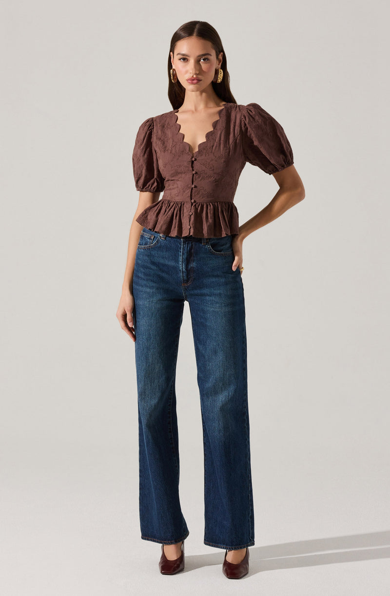 A woman is wearing a brown, textured blouse with puffed short sleeves and a scalloped V-neckline. The blouse has a button-up front with a fitted waist and a peplum hem. She pairs it with high-waisted, straight-leg blue jeans and brown heels.