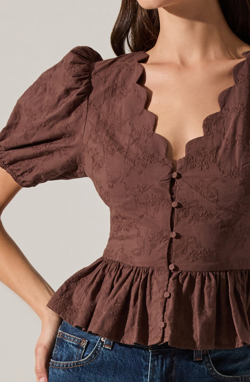 A close-up of a woman wearing a brown, textured, short-sleeved blouse with puffed sleeves. The blouse features a deep, scalloped neckline and a row of fabric-covered buttons down the front. The waistline is gathered with a peplum-style ruffle.