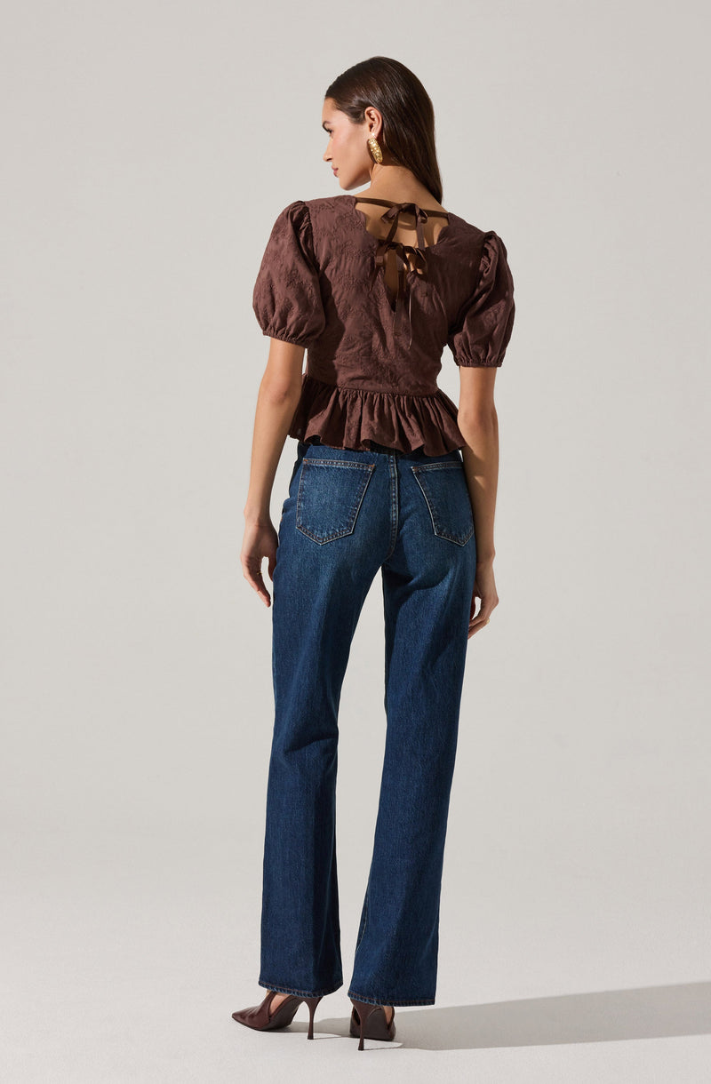 A woman is wearing a dark brown satin blouse with puffed short sleeves and a peplum hem. The back of the blouse features a tied ribbon detail at the neckline and a sheer panel. She is also wearing high-waisted, straight-leg blue jeans with visible stitching and a classic five-pocket design. Her footwear includes pointed-toe, high-heeled shoes.