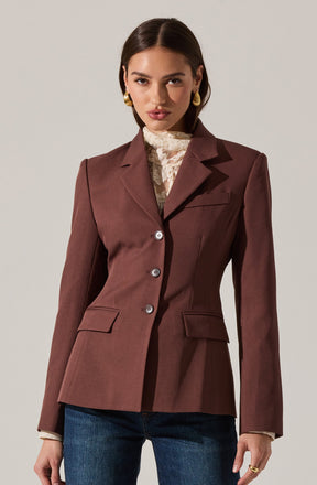 A woman is wearing a tailored, dark brown blazer with a structured fit, notch lapels, and three visible buttons on the front. She has on a high-neck, lace blouse underneath that is cream-colored with delicate floral patterns. She pairs the blazer with dark blue jeans and accessorizes with large, gold hoop earrings and a gold ring on her right hand.