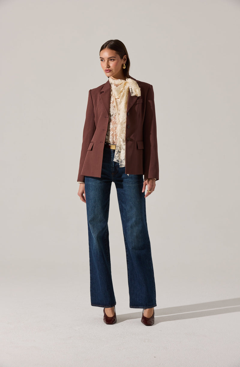A woman is wearing a tailored, dark brown blazer with notch lapels and a button closure. Underneath, she has a cream-colored lace blouse with ruffled detailing around the neckline and shoulders. She pairs this with high-waisted, straight-leg blue jeans and dark brown high-heeled shoes. She accessorizes with gold hoop earrings and a gold ring.