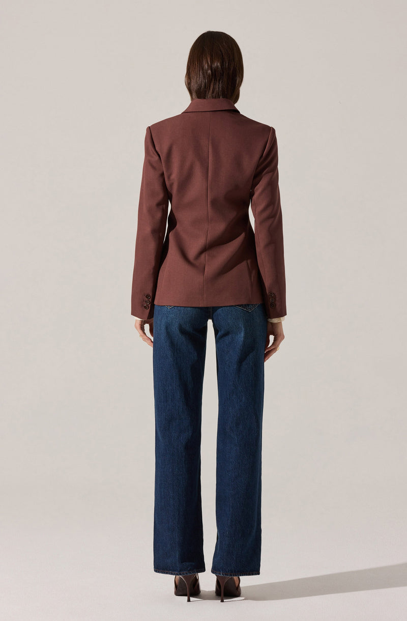 A person is wearing a tailored, brown blazer with a classic collar and long sleeves that have button details at the cuffs. They are also dressed in high-waisted, straight-leg blue jeans and brown high-heeled shoes.