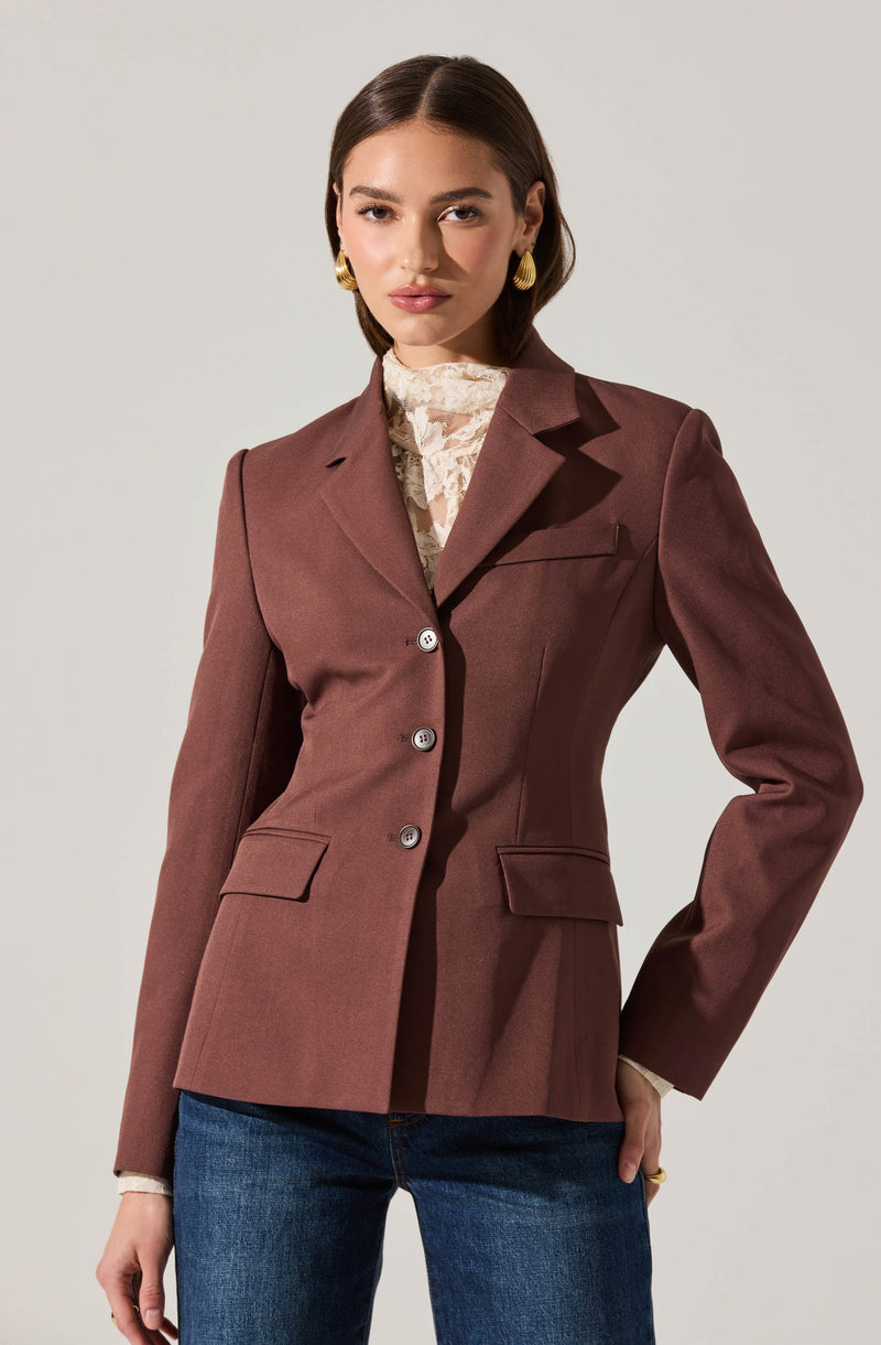A woman is wearing a fitted, single-breasted blazer in a muted mauve color. The blazer features notched lapels, four buttons down the front, and two flap pockets at the waist. Underneath, she has on a high-neck lace blouse in a light cream or beige color. She is also wearing medium-wash blue jeans.