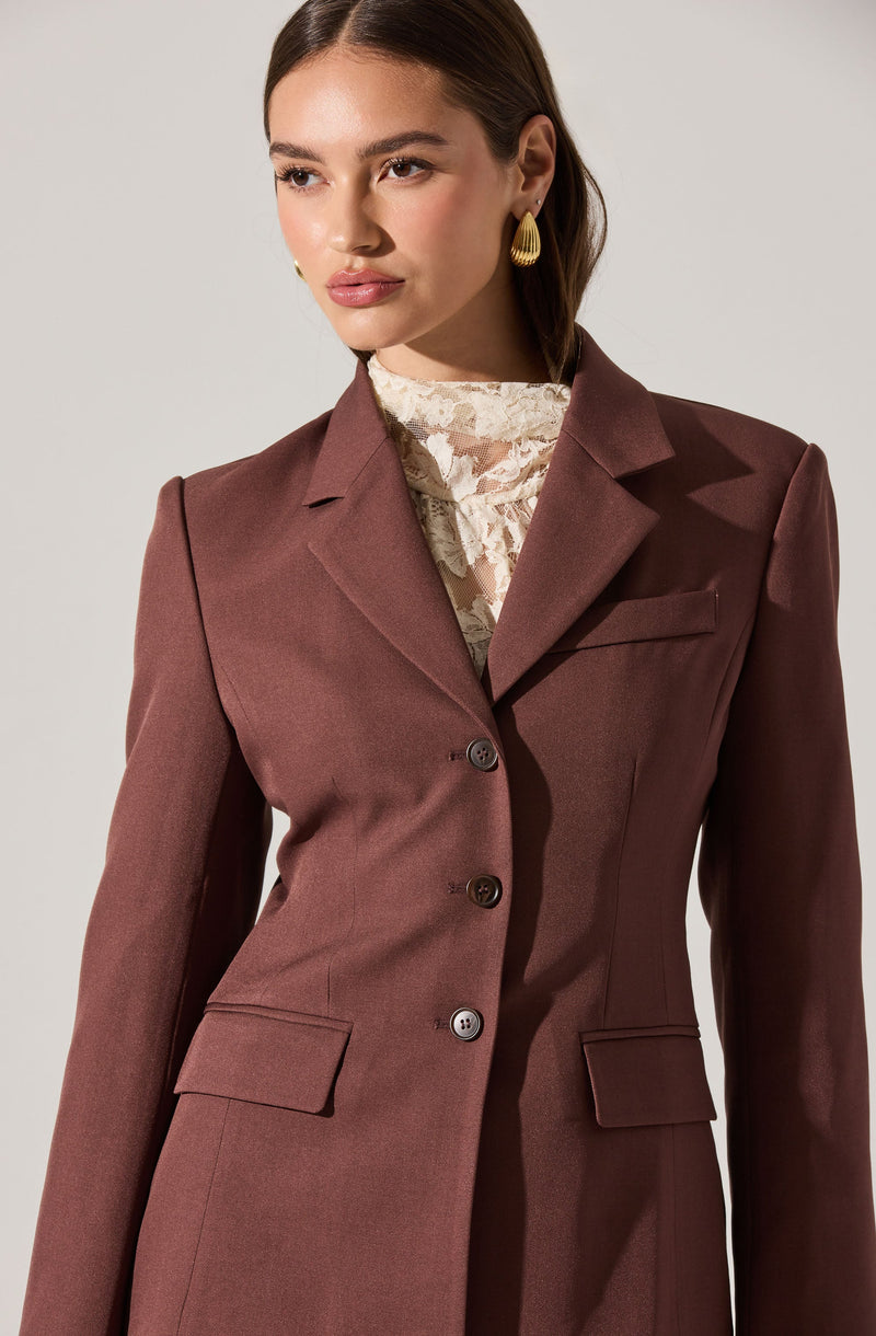 A woman is wearing a tailored, dark brown blazer with a notched lapel, three buttons down the front, and flap pockets on each side. Underneath, she has on a high-neck lace blouse in a light, cream color. She is also wearing gold teardrop-shaped earrings.