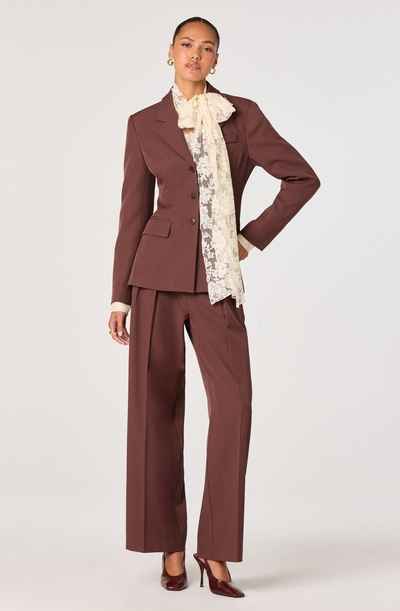 A woman is wearing a tailored, brown pantsuit consisting of a fitted blazer with structured shoulders and matching wide-leg trousers. She has a cream-colored lace blouse underneath that features a high neckline and a large bow tie at the collar. Her accessories include small hoop earrings, and she is wearing pointed-toe high heels in a matching brown shade.