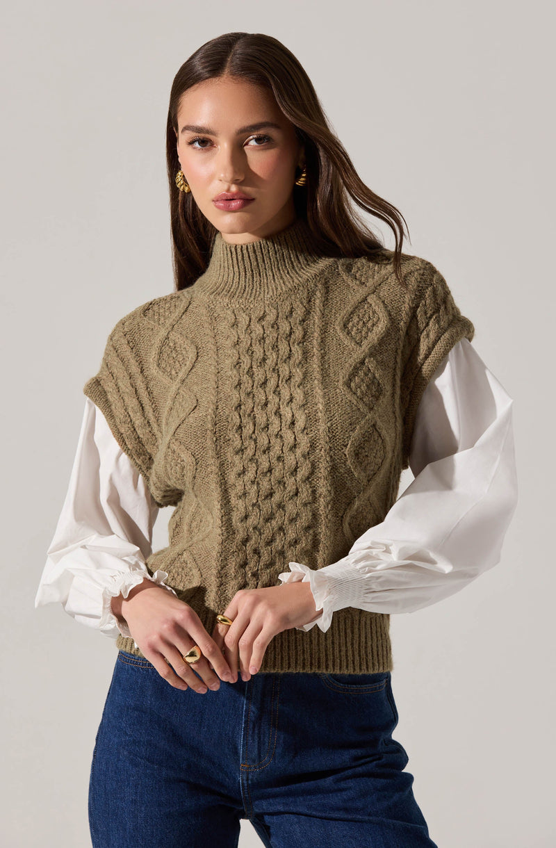 A woman is wearing a sleeveless, khaki-colored knitted sweater vest with textured cable and diamond patterns over a white blouse with puffy, gathered long sleeves. She is also wearing dark blue jeans and gold earrings.