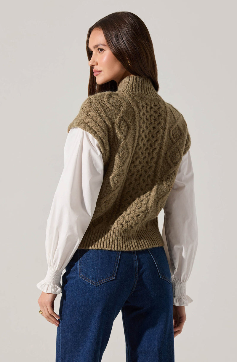 A woman is wearing a chunky, sleeveless, olive-green cable-knit sweater vest over a white blouse with long, slightly puffy sleeves and elasticized cuffs. She is also wearing dark blue jeans.