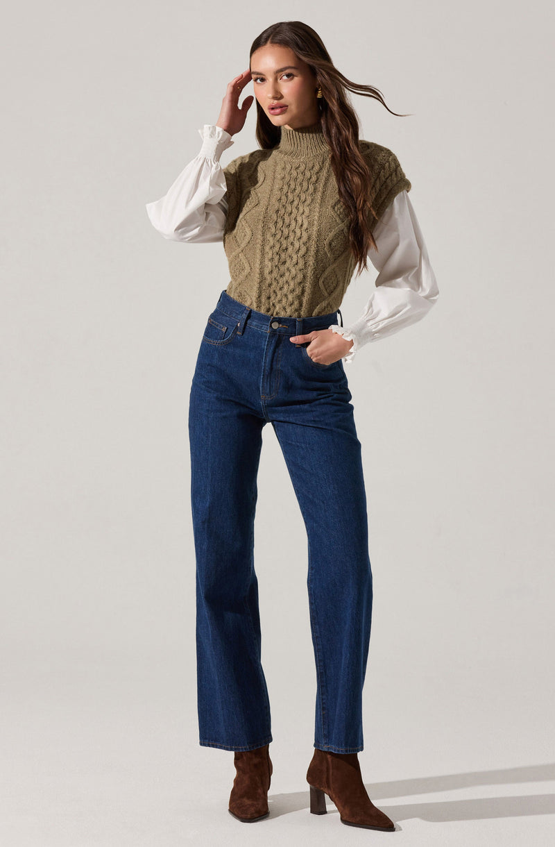 A woman is wearing a high-neck, sleeveless, olive green cable-knit sweater vest layered over a white blouse with long, puffy sleeves and ruffled cuffs. She pairs the top with high-waisted, straight-leg blue jeans and brown suede ankle boots with a chunky heel.