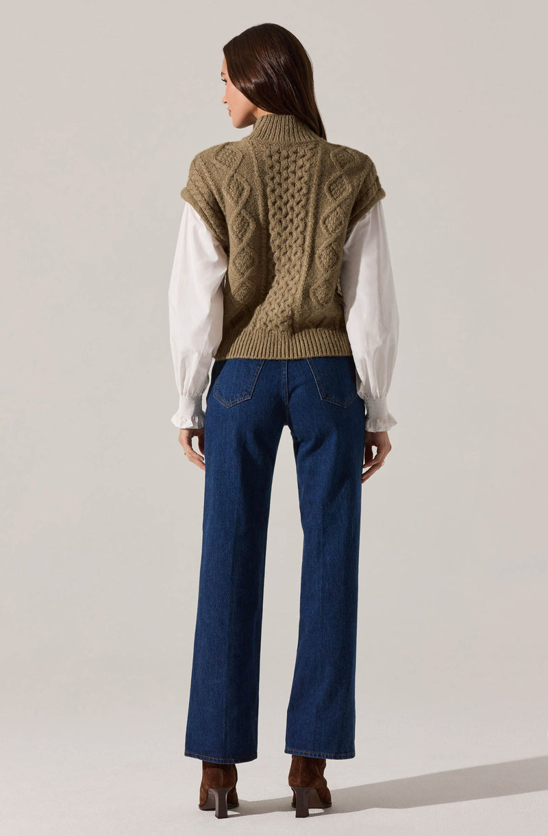 A woman is wearing a chunky knit sleeveless sweater vest in a tan color, featuring a textured cable knit pattern. Underneath, she has on a long-sleeve white blouse with gathered cuffs. She is also wearing high-waisted, straight-leg blue jeans and brown heeled ankle boots.