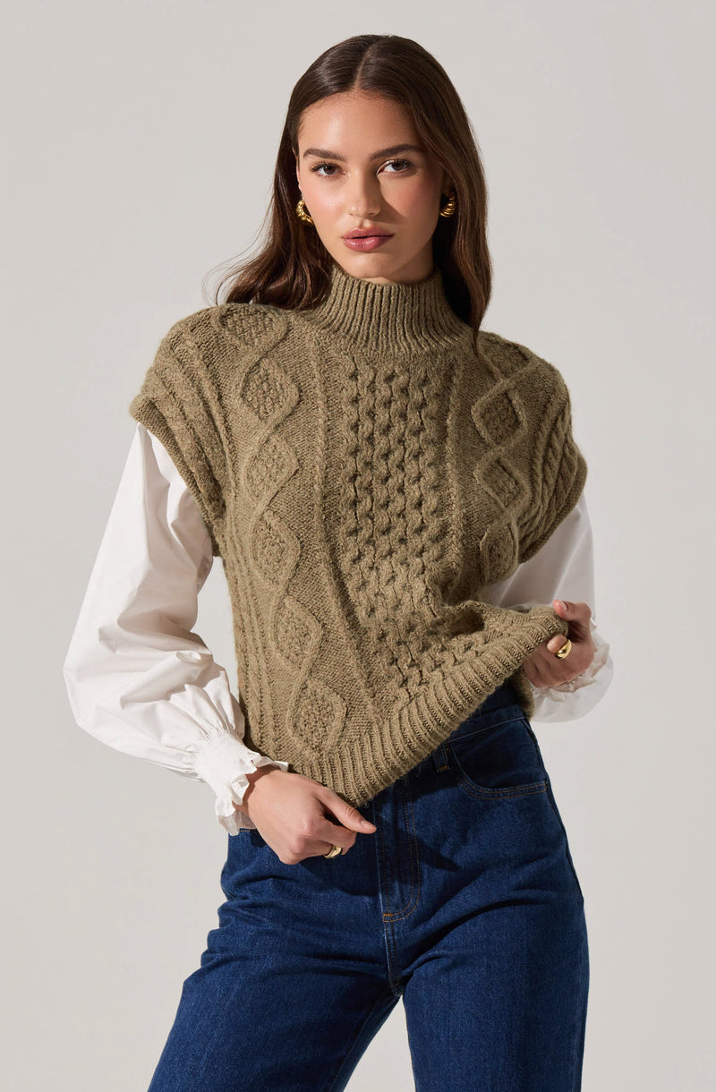 A woman is wearing a chunky, sleeveless, olive-green knit sweater with cable and diamond patterns. Underneath, she has on a white long-sleeve blouse with puffed, gathered cuffs. She pairs the outfit with high-waisted blue jeans and accessorizes with gold hoop earrings.