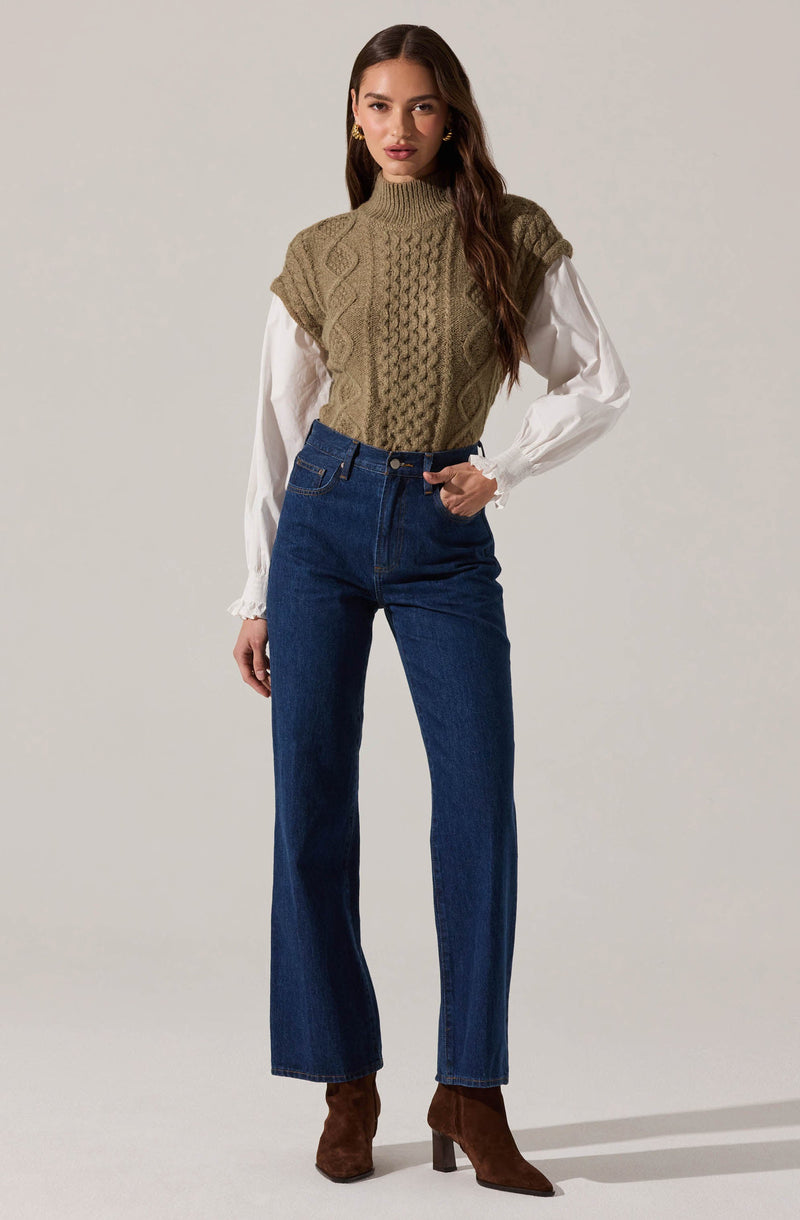 A woman is wearing a high-neck, sleeveless, knitted vest in an olive green color with cable-knit patterns. Underneath, she has on a long-sleeved white blouse with ruffled cuffs. She is also wearing high-waisted, dark blue straight-leg jeans and brown suede ankle boots with block heels.
