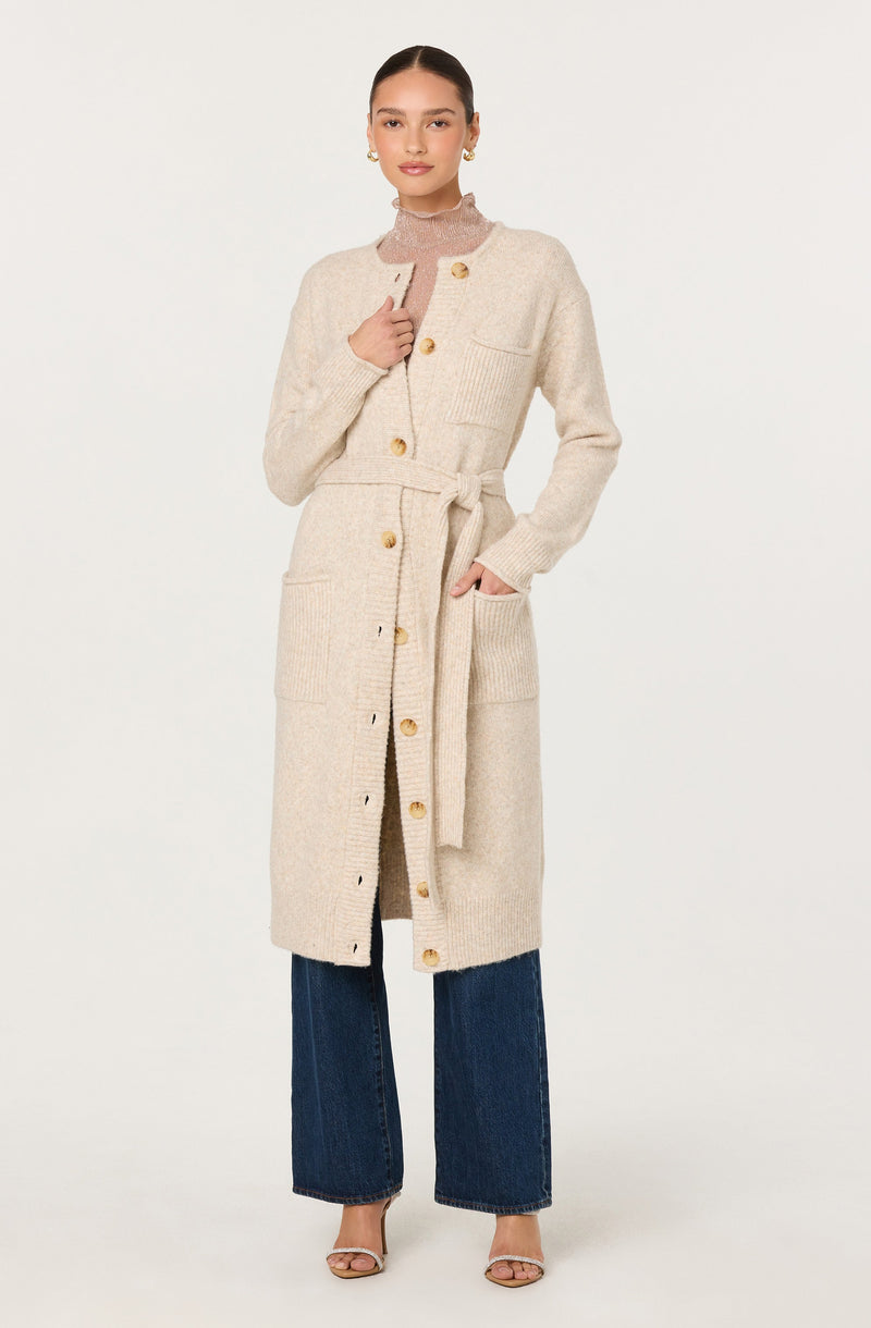 A woman is wearing a long, cream-colored knit coat with large buttons down the front and a belt tied at the waist. The coat features a chest pocket on the left side and large patch pockets on the hips. Underneath, she has on a sheer, high-neck top, and she is also wearing wide-leg blue jeans. She has on open-toe heels and gold hoop earrings.