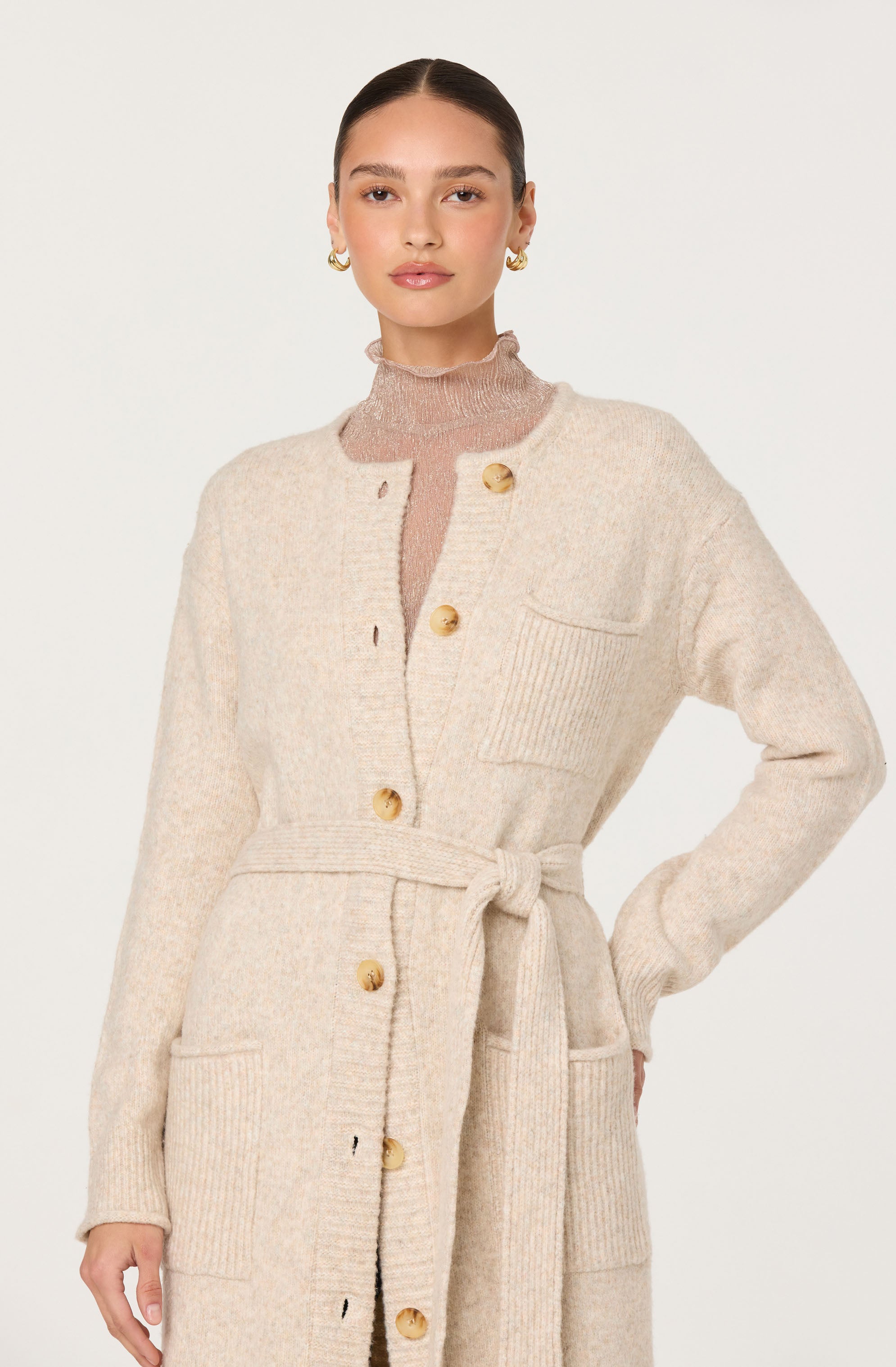 A woman is wearing a long, cream-colored knit cardigan with large front pockets and gold buttons down the front. The cardigan features a matching tied belt at the waist. Underneath, she has on a light pink, semi-sheer, high-neck sweater. She is accessorized with gold hoop earrings.