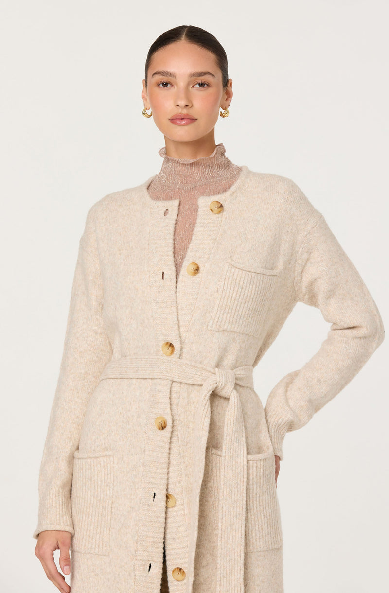 A woman is wearing a long, cream-colored knit cardigan with large front pockets and gold buttons down the front. The cardigan features a matching tied belt at the waist. Underneath, she has on a light pink, semi-sheer, high-neck sweater. She is accessorized with gold hoop earrings.