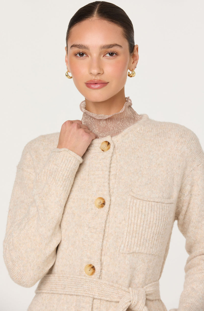 The woman is wearing a beige, textured knit cardigan with large, round, tortoise-shell buttons down the front and a matching waist tie. She has a sheer, light pink ruffled turtleneck underneath the cardigan. She is also wearing gold hoop earrings.