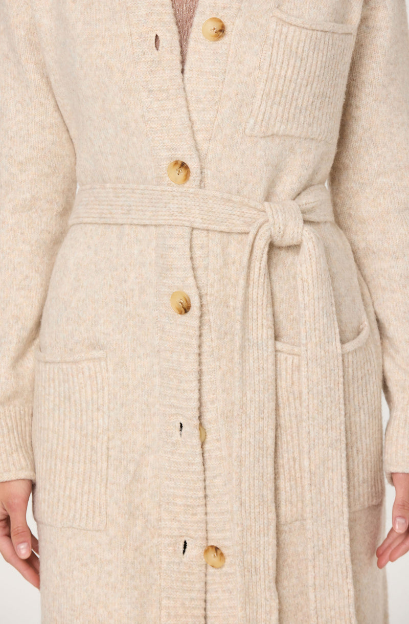 A long, cream-colored knit cardigan with large wooden buttons down the front. It has a ribbed texture at the cuffs, pockets, and waist, and features a matching knit belt tied at the waist. The cardigan appears to be made of a soft, cozy material.