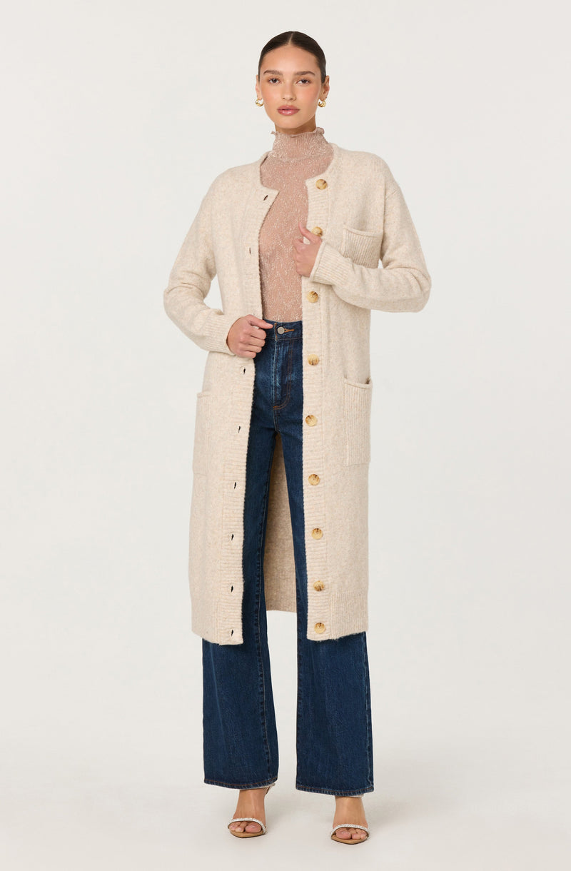 A woman is wearing a long, beige, textured knit cardigan with large yellow buttons down the front and a patch pocket on her left side. Underneath, she has on a light pink, fitted turtleneck top. She pairs the outfit with high-waisted, dark blue wide-leg jeans and nude-colored open-toe heels.