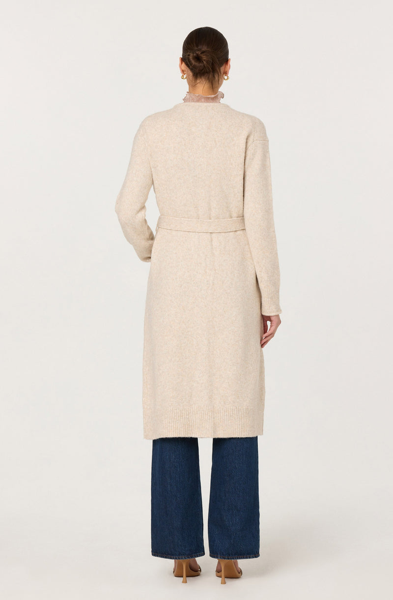 A woman is wearing a long, cream-colored, knitted cardigan that reaches below her knees. The cardigan has a belt at the waist and a ribbed hem and cuffs. She is also wearing wide-leg dark blue jeans and beige high-heeled shoes.