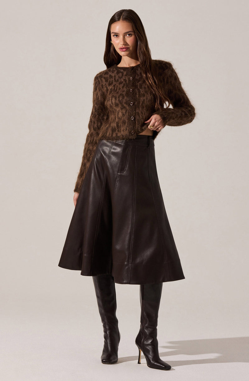 A woman is wearing a long-sleeved, brown fuzzy cardigan with a leopard print pattern and buttons down the front. She pairs it with a high-waisted, A-line leather skirt in dark brown or black that falls below the knees. She completes the outfit with tall, black leather high-heeled boots.