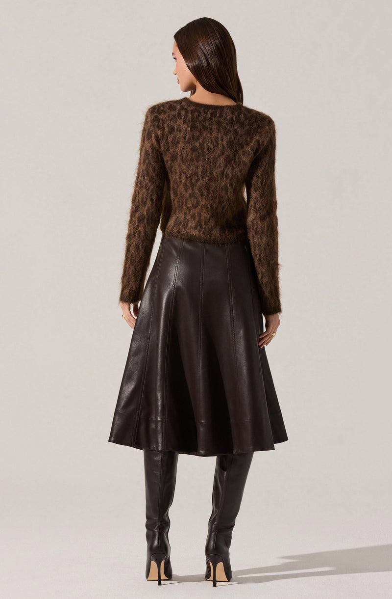 A woman is wearing a long-sleeved, brown, fuzzy sweater with a leopard print pattern and a high, round neckline. She pairs it with a high-waisted, dark brown leather A-line skirt that falls below the knees. She completes the outfit with tall, matching dark brown leather high-heeled boots.