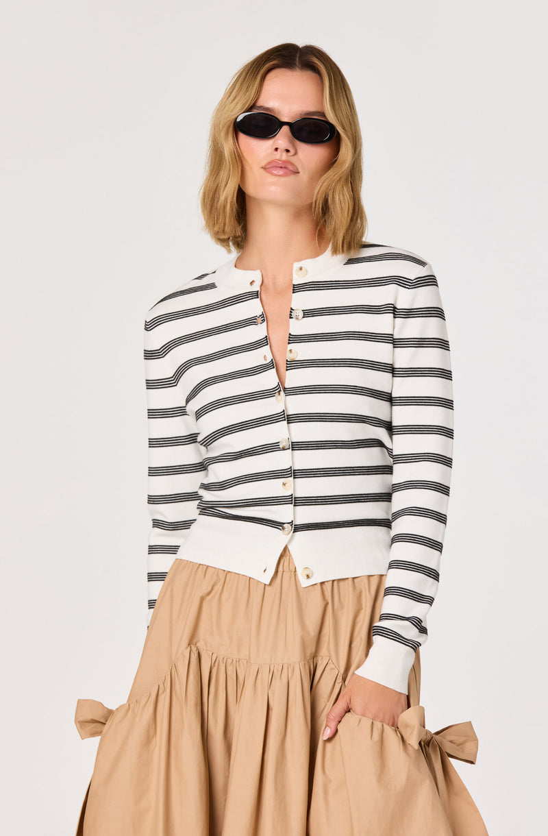 Woman wearing a white and black striped cardigan with beige skirt and black sunglasses.