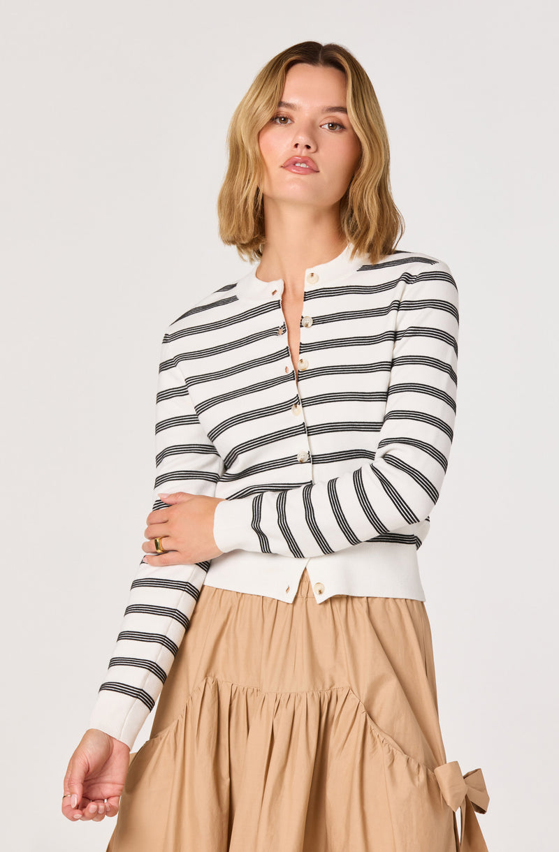 Woman wearing a white cardigan with black horizontal stripes and beige skirt with a bow detail.