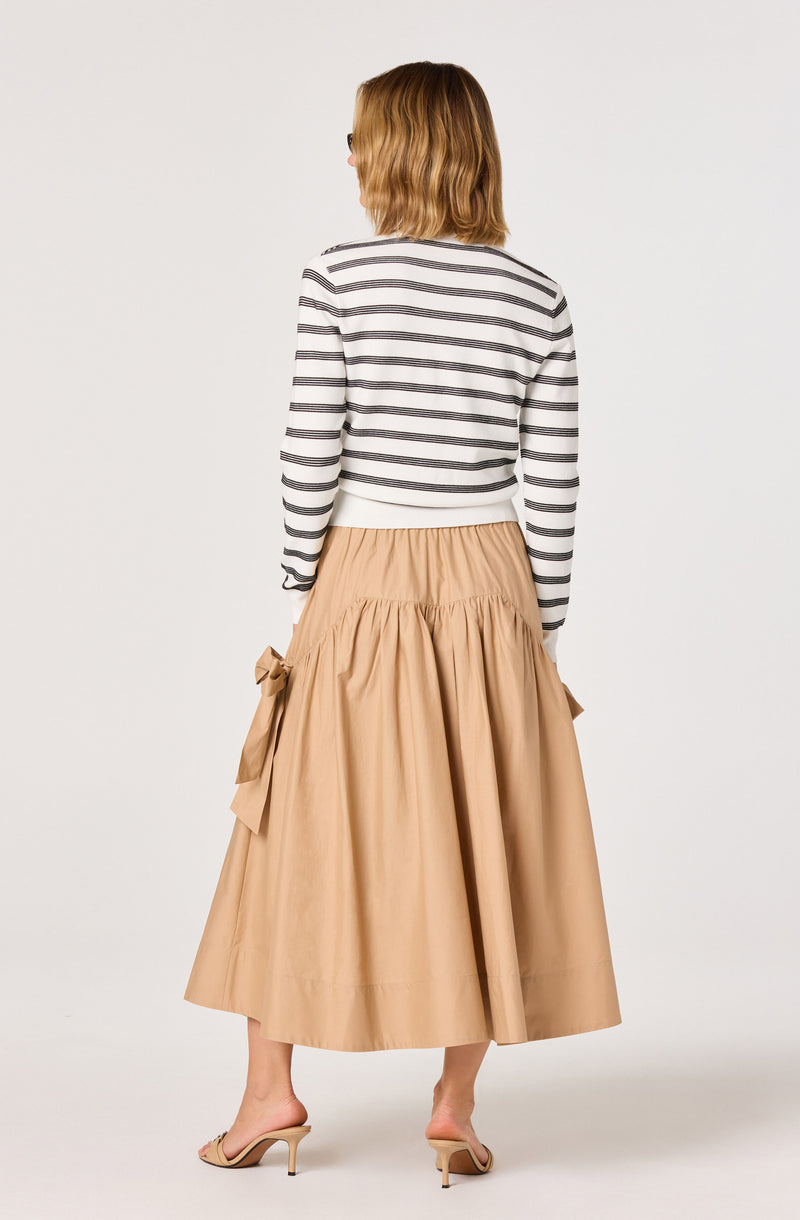 Woman wearing a white and black striped long-sleeve shirt and a tan, full, gathered midi skirt with tied bows.
