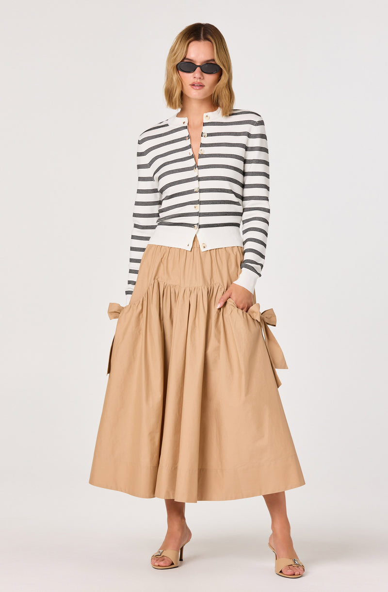 Woman wearing a white and black striped jacket, beige wide-leg skirt with bows, beige heels, and black sunglasses.