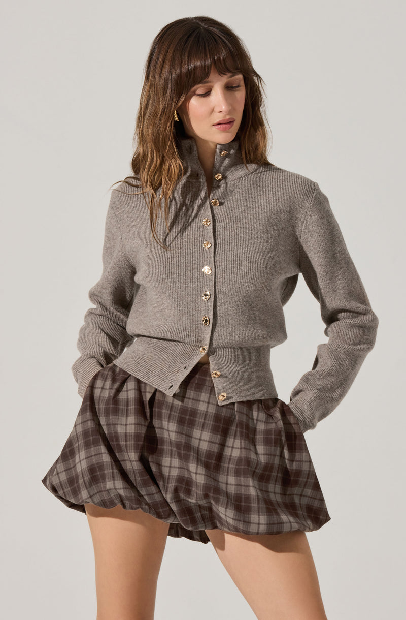 A woman is wearing a gray, long-sleeved, button-up cardigan with a high collar and gold buttons. She pairs it with loose-fitting, high-waisted plaid shorts in shades of brown and gray.