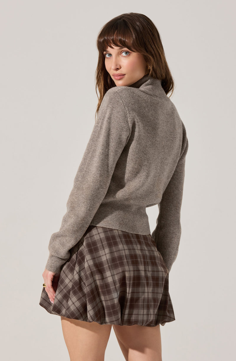 A woman is wearing a light gray, long-sleeved, turtleneck sweater with a slightly fitted waist. She pairs it with a brown plaid skirt that has a pleated design.